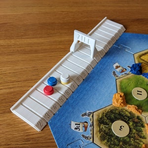 Op de afbeelding: Een wit 3D-geprint spelstuk met een genummerde baan en een kleine brug, ontworpen voor een bordspel. Het stuk is geplaatst op een blauw en groen spelbord met diverse spelstukken.