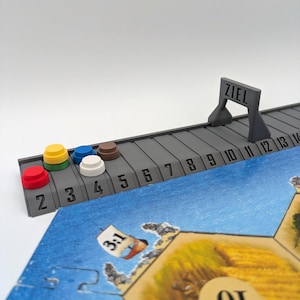 Könnte beinhalten: Ein Brettspiel-Setup mit einer grauen Strecke mit Zahlen und dem Wort "ZIEL". Bunte Spielsteine ​​liegen auf der Strecke. Ein Spielbrett mit einer blau-beigen Landschaft befindet sich im Vordergrund. Das Brett hat die Nummer "10".