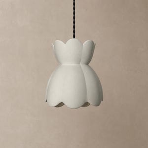 May include: A matte white pendant light fixture with a scalloped, flower-like silhouette. The shade features a smooth, curved design that flares outward at the bottom and is suspended from a black twisted fabric cord extending from the top.