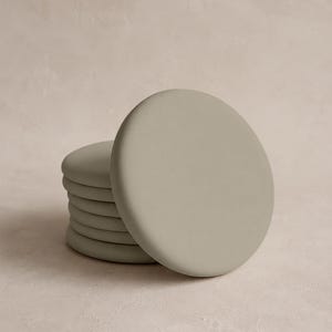 May include: A stack of six circular coasters in a solid matte sage green color. Each coaster features a smooth, rounded edge and a flat surface, arranged in a slightly offset vertical pile to show the thickness of each individual piece.