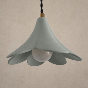 May include: A light blue, flower-shaped pendant light fixture with a white bulb. The fixture has a brass-colored top and a black twisted cord. The light is hanging against a neutral background.