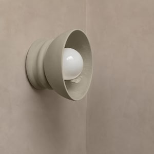 May include: A wall-mounted light fixture featuring a matte, light beige, funnel-shaped ceramic base with a smooth, rounded profile. A single, spherical white glass light bulb is centered within the wide, open aperture of the ceramic housing.