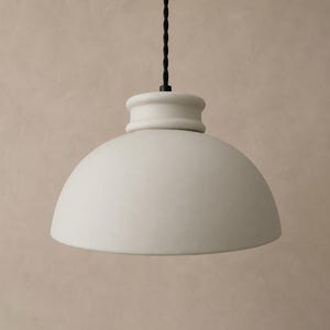 May include: A matte off-white pendant light featuring a smooth, dome-shaped shade with a rounded top collar. The fixture is suspended by a thin, black twisted fabric cord that connects to the top center of the lamp housing.