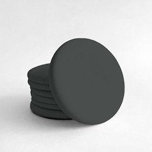 May include: A stack of six dark gray, round coasters. One coaster is leaning against the stack. The coasters are made of a solid, matte material. The background is plain white.