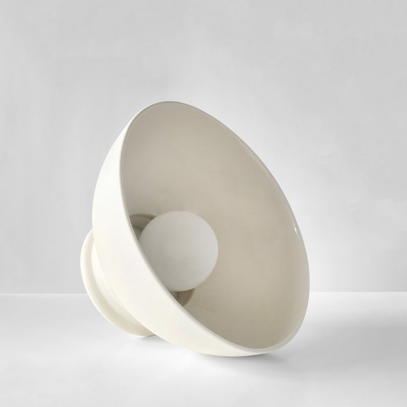 Op de afbeelding: Een witte keramische lamp met een schaalvormige kap en een gloeilamp erin.