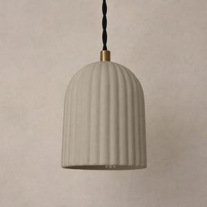 May include: A cream-colored, ribbed pendant light fixture with a rounded, bell-like shape. The light hangs from a black cord with a gold-colored metal accent at the top. The light fixture is a neutral color and has a minimalist design.