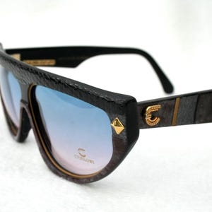 Könnte beinhalten: Sonnenbrille mit schwarzem, texturiertem Rahmen und blau verlaufenden Gläsern. Die Bügel sind mit einem goldenen Logo-Emblem und einem pyramidenförmigen Nietendetail versehen. Das Glas trägt ein goldenes Logo. Der Rahmen hat einen markanten Steg.