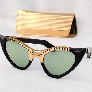 May include: Cat-eye sunglasses featuring black frames with gold-tone metal accents along the brow line, light green tinted lenses, and a floral engraving at the bridge. A matching gold-tone metallic hard-shell glasses case is positioned behind.