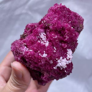 May include: A vibrant magenta mineral specimen with a rough, crystalline texture. The rock features deep pink, porous surfaces interspersed with clusters of small, reflective, light-catching crystals that create a sparkling effect.