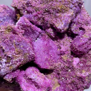 May include: Close-up of a cluster of vibrant purple mineral formations. The crystalline structures have a rough, textured surface with hints of gold and brown. The overall appearance is a rich, saturated color, showcasing natural geological beauty.