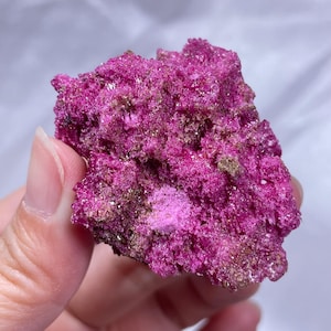 May include: A raw mineral specimen featuring a vibrant, saturated magenta and fuchsia crystalline structure. The surface is highly textured with small, sparkling points and interspersed patches of metallic bronze and gold-toned mineral deposits.