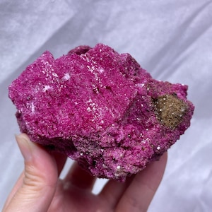 May include: A raw, vibrant magenta mineral specimen with a crystalline, textured surface. The rock features deep pink hues, small sparkling facets, and a small patch of brownish, earthy material on the right side of the irregular shape.