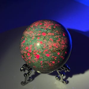 May include: A green and red orb, possibly a gemstone, sits on a decorative silver stand. The orb has a speckled pattern of red spots against a green background. The stand has a detailed, ornate design.