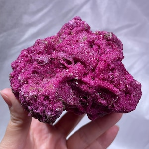 May include: A vibrant magenta mineral specimen with a porous, crystalline texture. The rock features deep crevices and a rough, jagged surface with small, sparkling points throughout its structure, showcasing a consistent, intense pinkish-purple hue.
