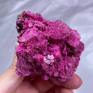 May include: A vibrant, deep magenta mineral specimen with a crystalline, textured surface. The rock features jagged, uneven edges and small, sparkling facets that catch the light, displaying a consistent, intense pinkish-purple hue throughout.