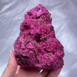 May include: A vibrant magenta mineral specimen with a rough, crystalline texture. The rock features deep crevices and a jagged, uneven surface with small, sparkling points throughout, showcasing a rich, saturated pinkish-purple hue.