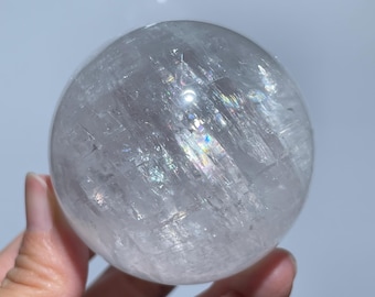 May include: A clear quartz crystal sphere. The sphere is round and polished, with internal fractures and iridescent rainbow reflections. The crystal is held in a hand against a soft, neutral background. This decorative crystal sphere is a unique home decor item.