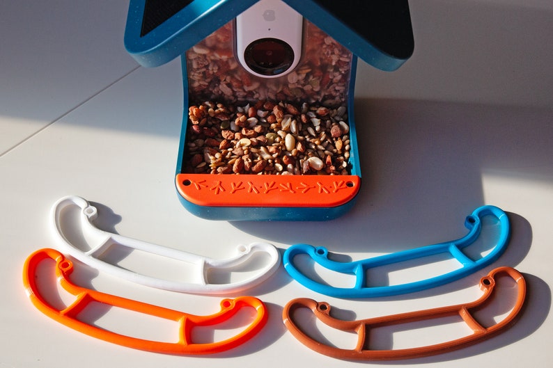 K&ouml;nnte beinhalten: Vier verschiedene farbige Vogel futterplatz Sitzstangen aus Kunststoff. Die Sitzstangen sind in Form eines Kanus und sind wei&szlig;, orange, blau und braun. Die Sitzstangen befinden sich auf einer wei&szlig;en Oberfl&auml;che.