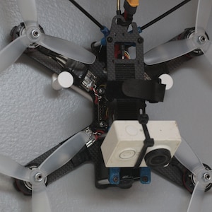 Puede incluir: Un dron blanco con cuatro hélices transparentes y una cámara blanca montada en la parte delantera. El dron está colgado del techo.