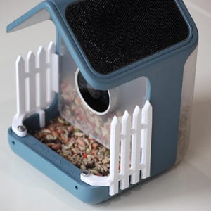 Könnte beinhalten: Ein moderner Vogelfutterautomat in Hausform mit blauem Rahmen, weißem Zaun und schwarzem Mesh-Dach. Der Futterautomat ist mit Vogelfutter gefüllt und hat eine transparente Kunststoffseite.