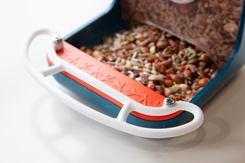 K&ouml;nnte beinhalten: Nahaufnahme eines blauen und orangen Vogelhauses mit einer wei&szlig;en Sitzstange. Der Futterspender ist mit verschiedenen Vogelfutter gef&uuml;llt.