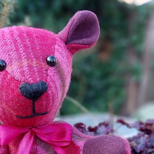 May include: A pink and brown plaid teddy bear with black eyes and a pink bow. The bear is sitting on a wooden surface.