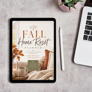 May include: A digital tablet displaying a Fall Home Reset Planner cover with text, a cozy living room scene featuring a mug, a candle, and textured pillows. A white stylus rests beside the tablet, which is positioned next to a laptop computer.