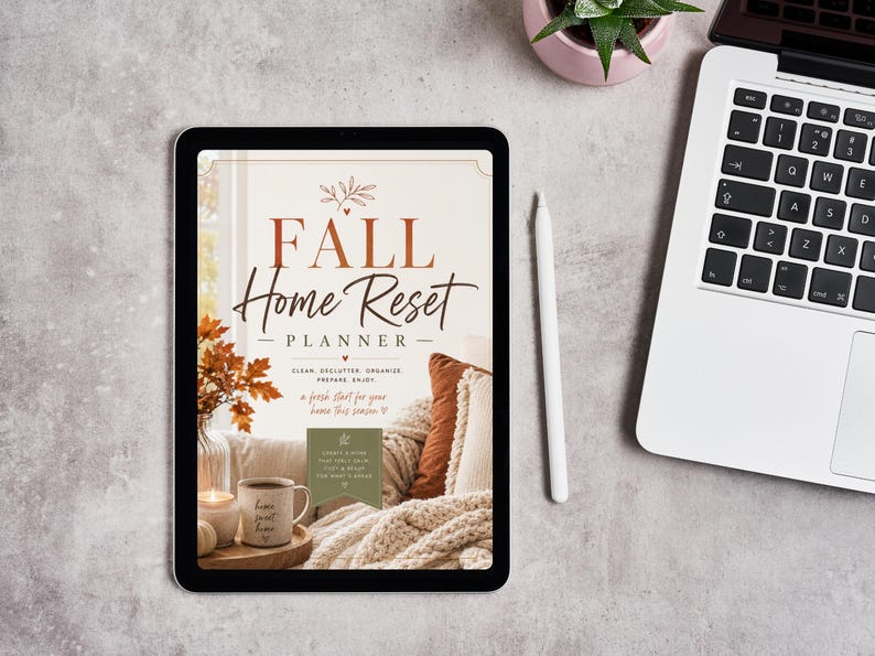 May include: A digital tablet displaying a Fall Home Reset Planner cover with text, a cozy living room scene featuring a mug, a candle, and textured pillows. A white stylus rests beside the tablet, which is positioned next to a laptop computer.