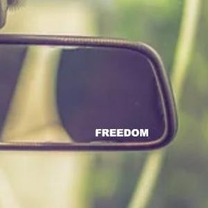 May include: A rectangular rearview mirror with a dark reflective surface. The word "FREEDOM" is printed in white across the mirror's surface. The mirror is framed in a dark color, and the background is blurred.