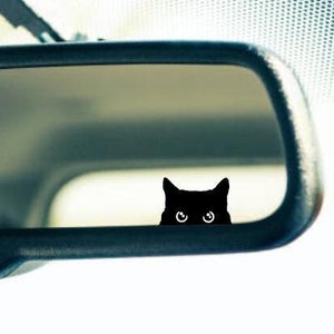 May include: A black cat sticker peeking out from behind a rearview mirror in a car.