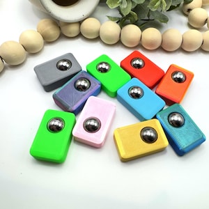 May include: A collection of colorful, rectangular fidget toys, each with a silver ball bearing in the center. The toys are in various colors, including green, pink, blue, and orange. The background is white.