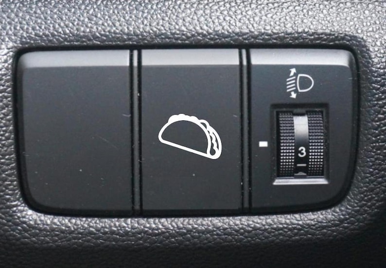 May include: A black car dashboard with three buttons. The left button is blank, the middle button has a white icon of a taco, and the right button has a knob with a number "3" and a white icon of a headlight with an arrow pointing up and down.