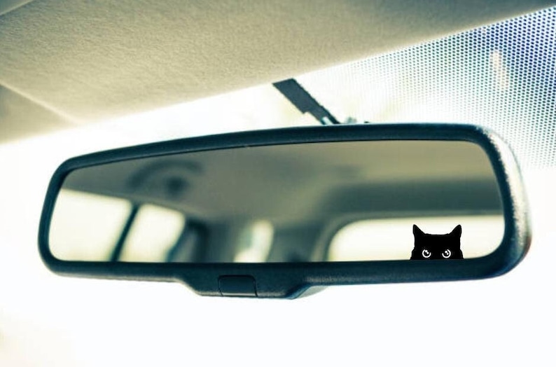 May include: A black cat sticker peeking out from behind a rearview mirror in a car.