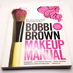 May include: A white book with a pink makeup brush and pink eyeshadow on the cover. The title is "BOBBI BROWN MAKEUP MANUAL" in black and pink lettering. The text "For Everyone from Beginner to Pro" is in black lettering. The text "Celebrating 20 Years of Making Women Beautiful" is in black lettering. The text "Indispensable...tons of foolproof beauty tips...includes everything. Bobbi Brown is an American makeup idol." -Glamour.com is in black lettering.