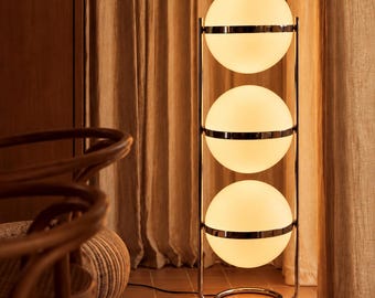 May include: A floor lamp featuring three stacked spherical white glass globes held within a vertical chrome metal frame. The lamp has a circular base and emits a warm, soft glow from each of the three illuminated glass spheres.