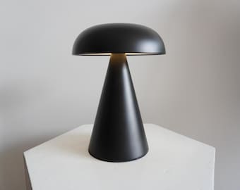May include: A black table lamp with a wide, rounded top and a conical base. The lamp is switched on and emitting a warm light.