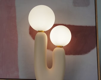 May include: A table lamp featuring a smooth, matte beige U-shaped base with two uneven vertical arms. Each arm is topped with a glowing, spherical white glass globe shade attached by a small gold-toned metal connector at the base of each bulb.