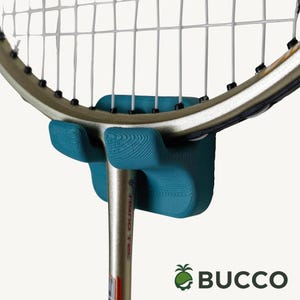 May include: A teal-colored, textured plastic badminton racket wall mount attached to the throat of a silver and black badminton racket. The mount features a grooved surface and is positioned to hold the racket frame securely. The text BUCCO is visible.