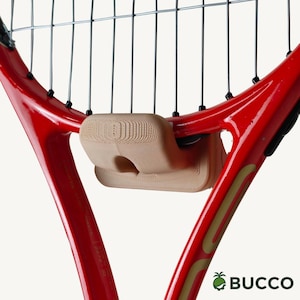 May include: A bright red tennis racket frame with a beige, textured, rectangular vibration dampener attached to the strings near the throat. The frame features gold accents and the text BUCCO with a small green pineapple icon in the bottom corner.