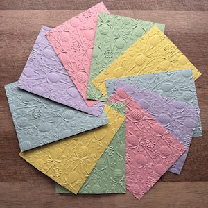 Peut inclure: Un ensemble de cartes en papier gaufré aux couleurs pastel, disposées en cercle. Chaque carte présente un motif répété d'œufs de Pâques, de fleurs et de nœuds. Les couleurs incluent le rose, le jaune, le vert et le lavande. Les cartes sont présentées sur une surface en bois.