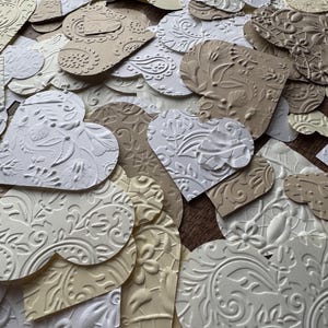 May include: A pile of white and beige paper hearts with embossed patterns. The hearts are arranged in a random pattern and some are overlapping.