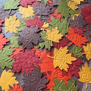 May include: A collection of colorful, textured paper leaves in shades of red, orange, yellow, green, and brown. The leaves have various embossed patterns, including honeycomb, swirls, and floral designs, creating a fall-themed decorative assortment.