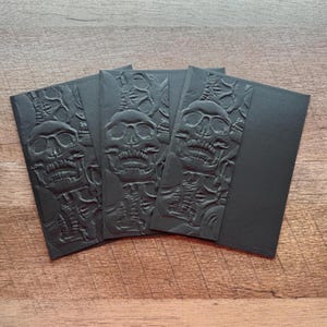 May include: Three rectangular black envelopes featuring a raised, embossed skull and skeletal pattern on the left side. The right side of each envelope is smooth and plain black. The design creates a monochromatic, textured appearance.