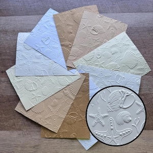 May include: A collection of rectangular paper sheets featuring embossed patterns of various seashells and starfish in shades of white, cream, and tan. A circular inset highlights the detailed texture of the raised shell and starfish designs.