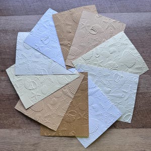 May include: A collection of embossed paper cards in various pastel colors, including white, beige, and light blue. Each card features a raised design of seashells and starfish, creating a textured, nautical theme. The cards are arranged in a circular pattern.