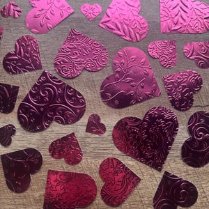 May include: A collection of various sizes of pink and magenta heart-shaped cutouts with embossed floral and swirl patterns. The hearts are scattered on a wooden surface, creating a decorative arrangement.