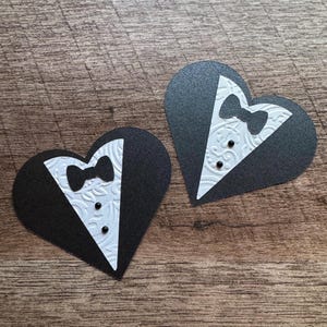 K&ouml;nnte beinhalten: Zwei schwarze herzf&ouml;rmige Papierschnitte mit wei&szlig;en Smoking-Designs. Jeder Ausschnitt zeigt eine wei&szlig;e Fliege und zwei schwarze Kn&ouml;pfe.