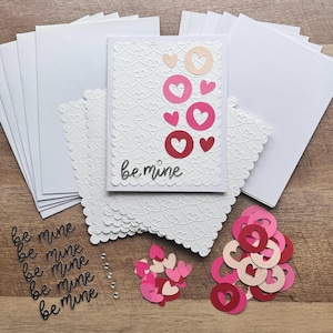 May include: A collection of Valentine's Day card-making supplies. Includes white cards, scalloped-edge cards, and a card with a heart design and the words "be mine." Also shown are heart-shaped cutouts and embellishments.