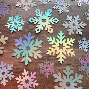 May include: Various sizes of iridescent snowflake cutouts. The snowflakes have a shimmering, multi-coloured finish, reflecting hues of blue, green, and purple. They are arranged on a wooden surface, creating a festive, wintery theme.