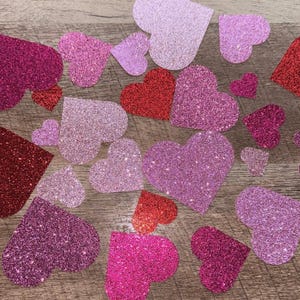 May include: A collection of pink, red, and purple glitter hearts. The hearts are scattered on a wooden surface.